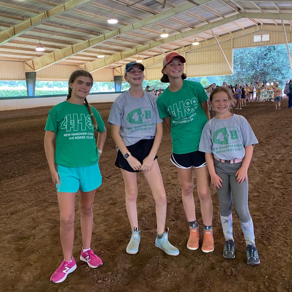 Four girls in 4-H tee shirts pose together in a covered riding ring.